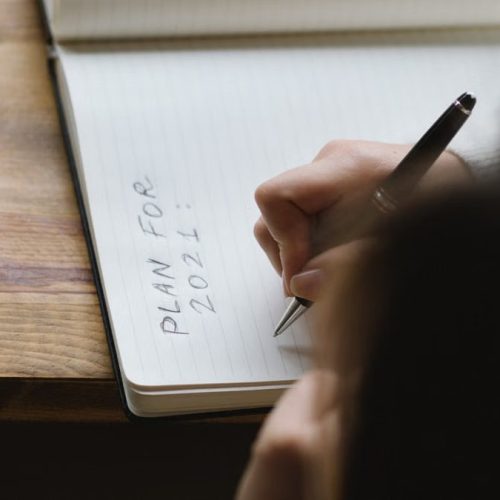 From above of crop unrecognizable female with dark hair sitting at wooden table and writing plan for 2021 in notebook