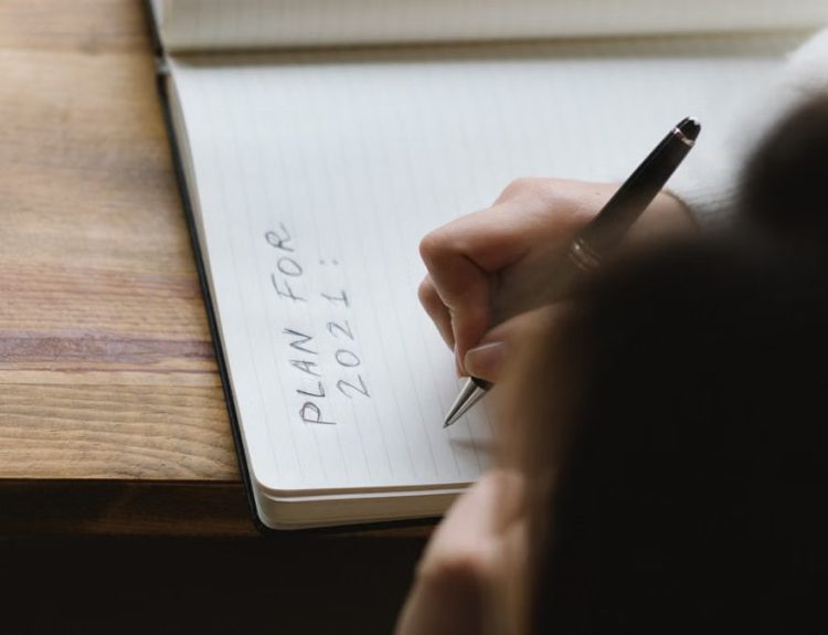 From above of crop unrecognizable female with dark hair sitting at wooden table and writing plan for 2021 in notebook